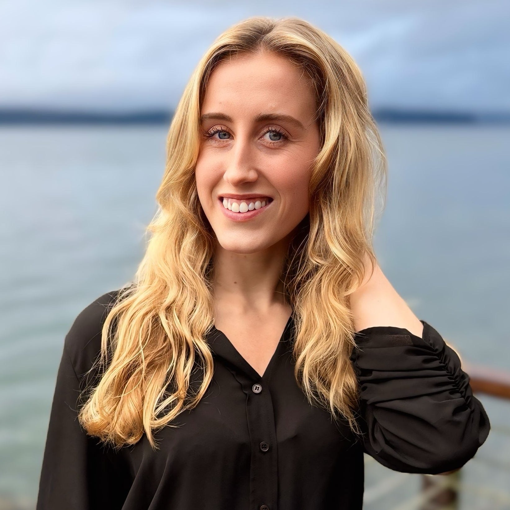 Woman standing by a body of water wearing a black blouse and brown skirt.