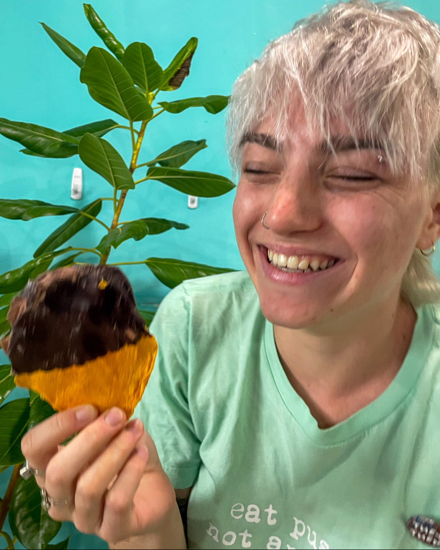 Smiling person holding a chocolate-dipped mango with a plant in the background