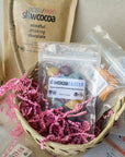slowcocoa craft chocolate Easter basket demonstration with various slowcocoa gift products including mindful drinking chocolate and creamer, pink crinkle paper, oatmilk chocolate bar on a light background