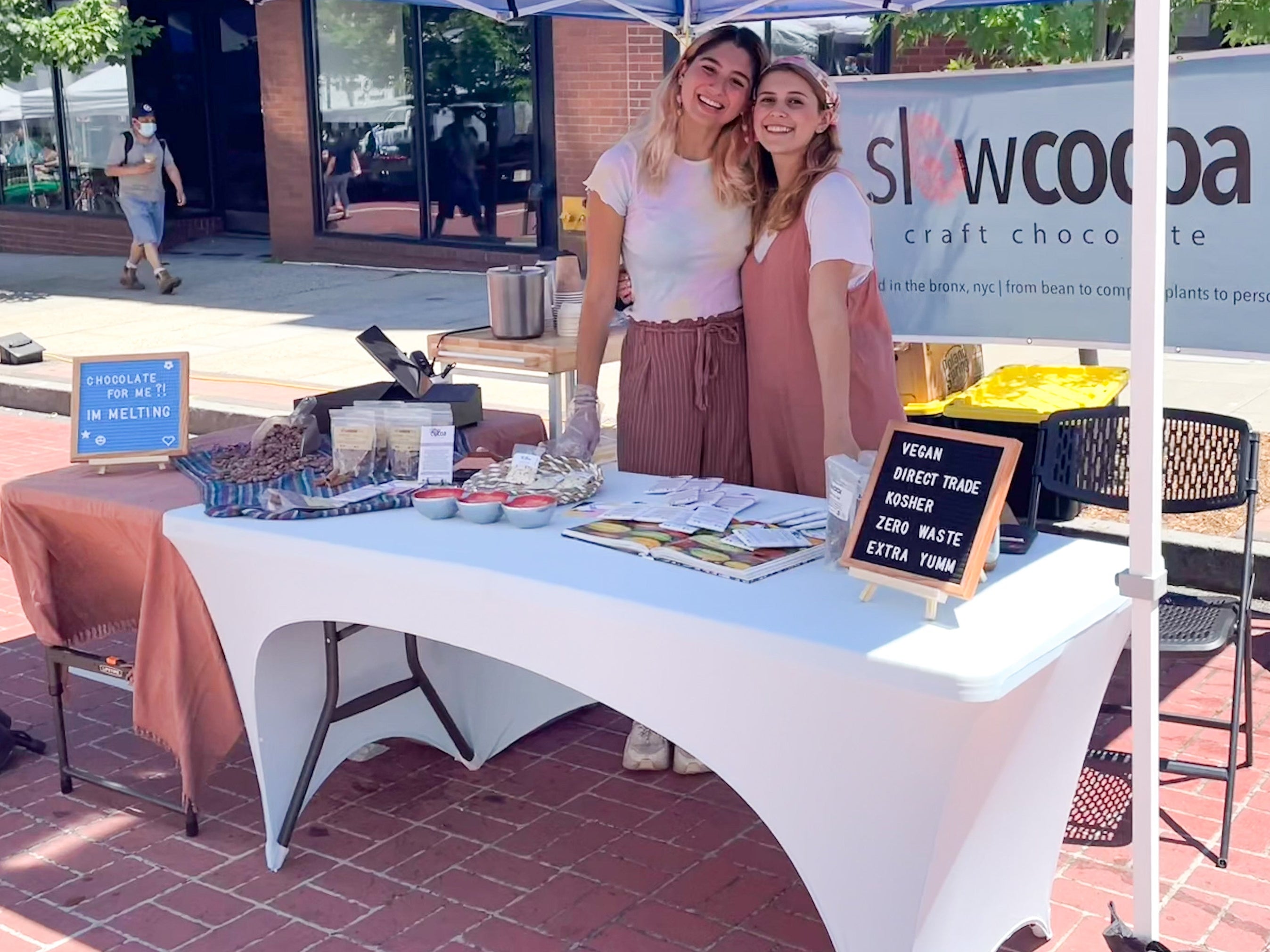 Two women standing under a blue tent with a table and promotional materials, including a sign for Slowcoa.