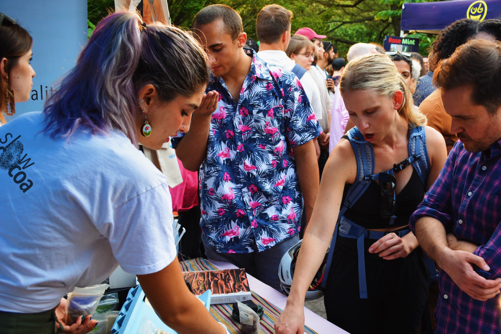 slowcocoa staff pointing to chocolate bars on the table at an outdoor crowded settings while several onlookers gather around the table