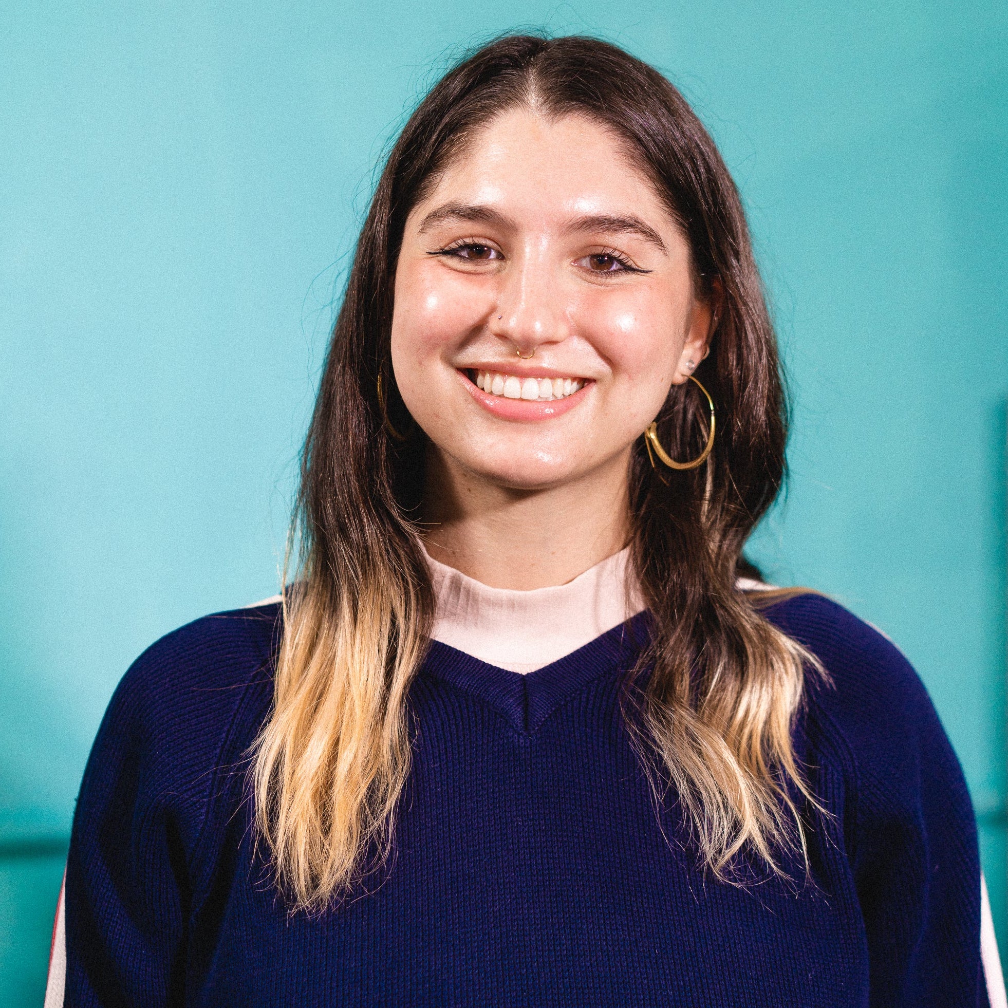 Smiling light-skinned woman with golden hooped earrings, long brown and highlighted hair, in a blue sweater