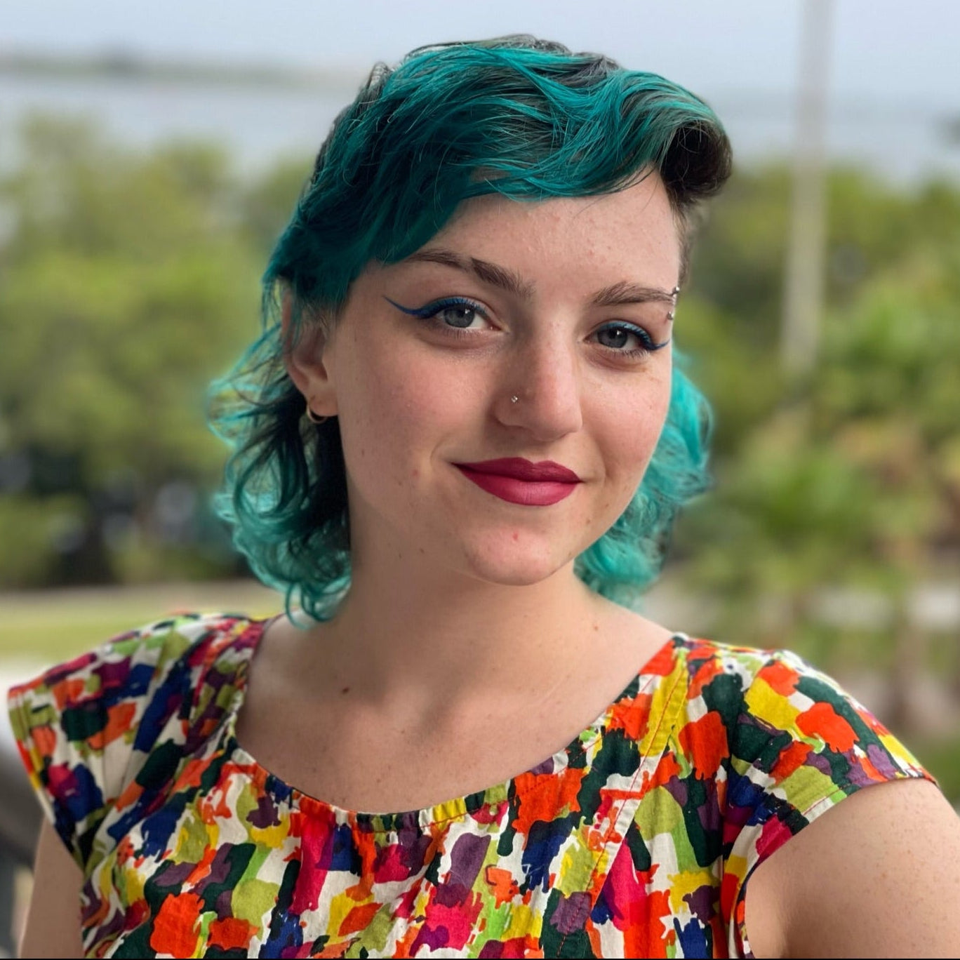 Person with teal hair and a colorful patterned dress standing outdoors with greenery in the background