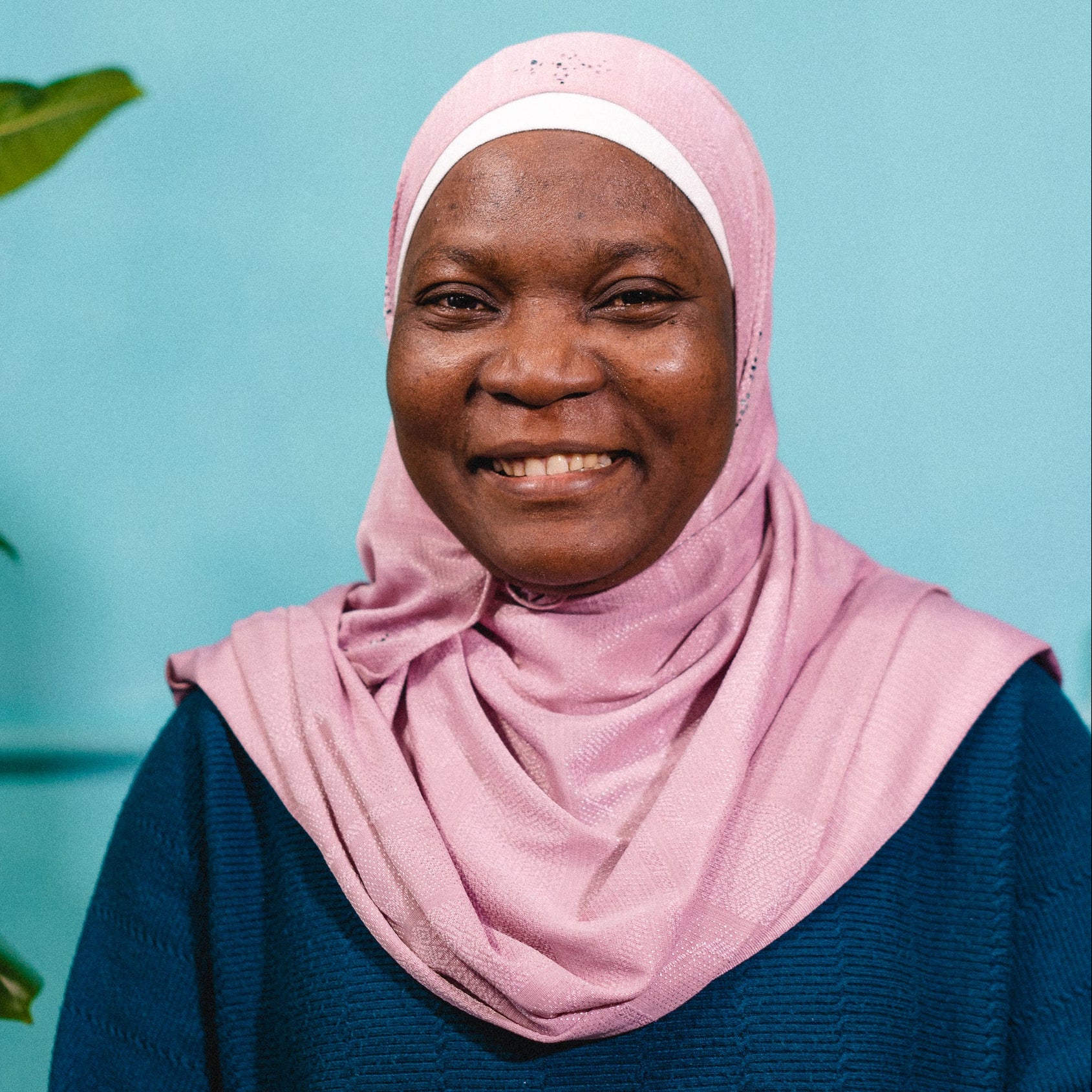 Woman wearing a pink hijab and blue top against a light blue background with a plant on the left.