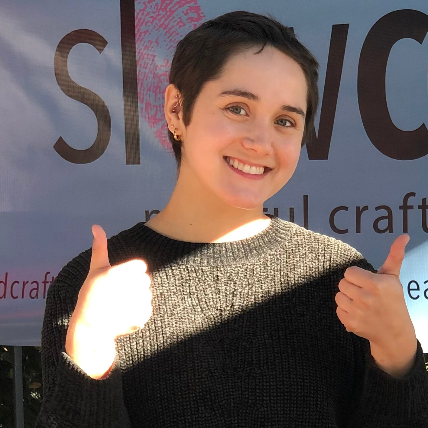 light skinned person smiling with thumbs up outdoors in front of a slowcocoa banner