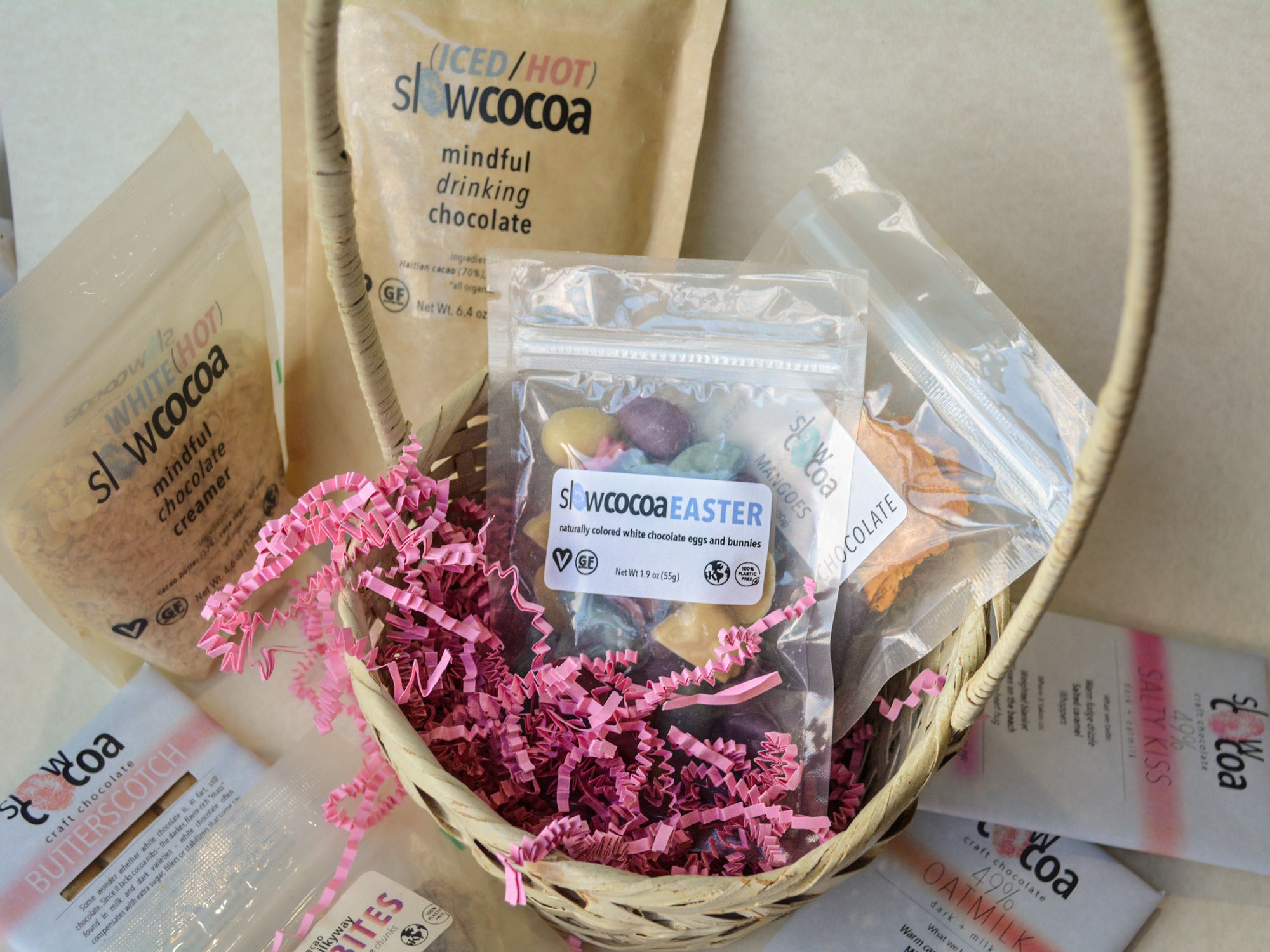 slowcocoa craft chocolate Easter basket demonstration with various slowcocoa gift products including mindful drinking chocolate and creamer, pink crinkle paper, oatmilk chocolate bar on a light background
