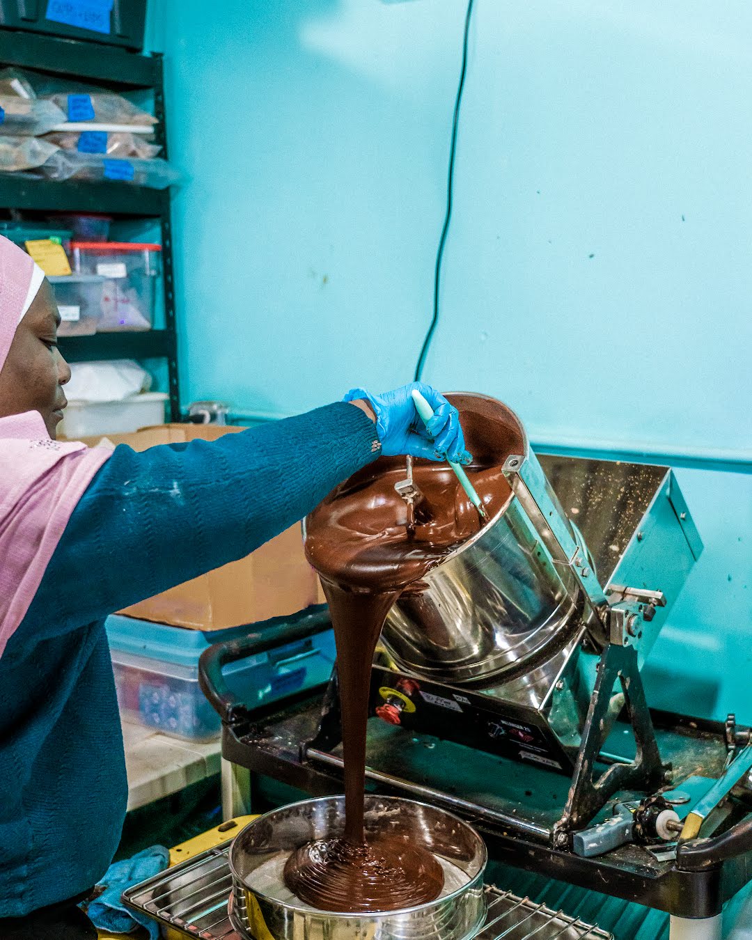 Slowcocoa production manager Zeinabou wearing a pink hijab, dark blue sweater, and blue gloves pours thick melted chocolate from a metal pot into a chocolate maker machine in a production setting.