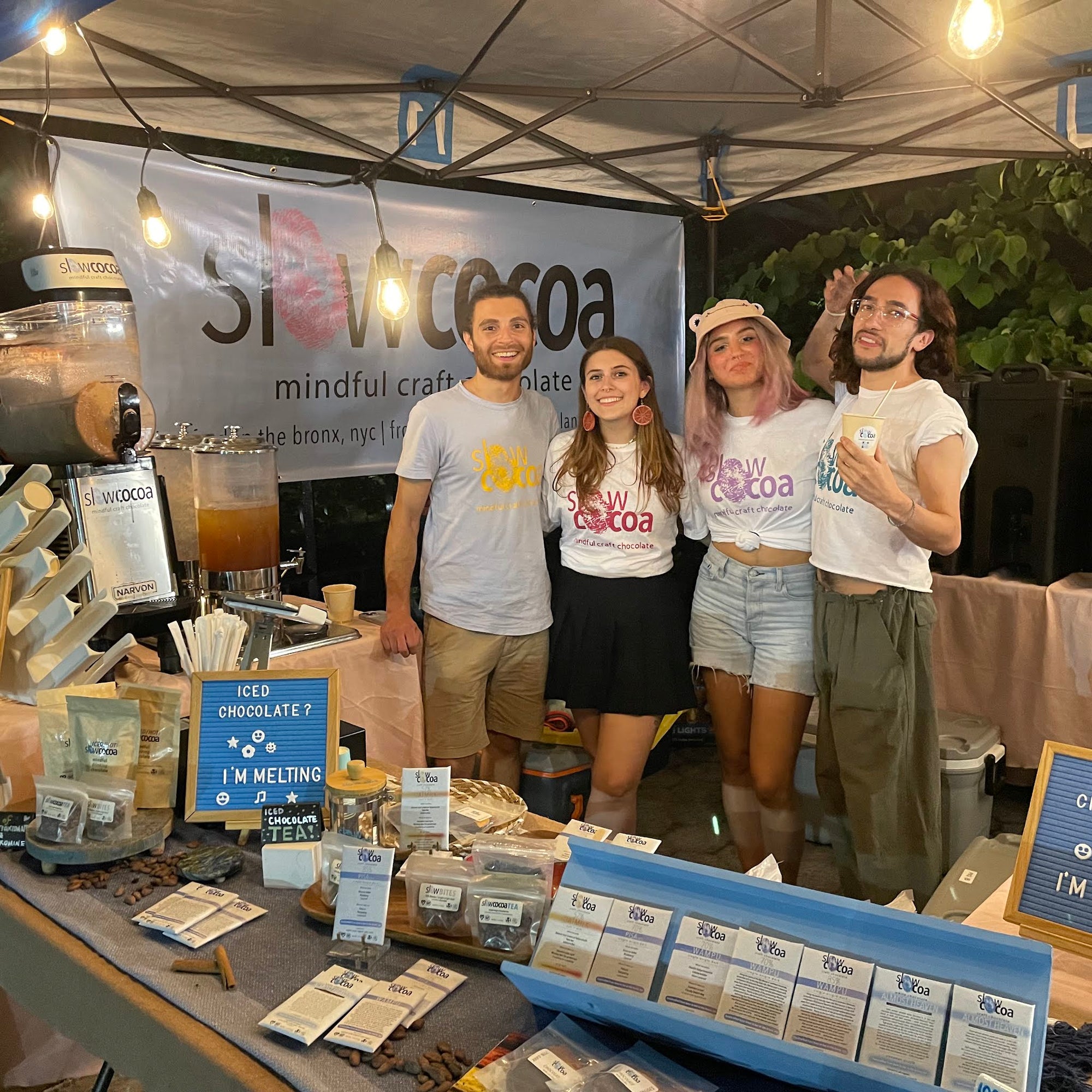 Four slowcocoa team members in branded tees behind the farmers market stall at night, with chocolate bars, cacao nibs and an iced chocolate dispenser on the table